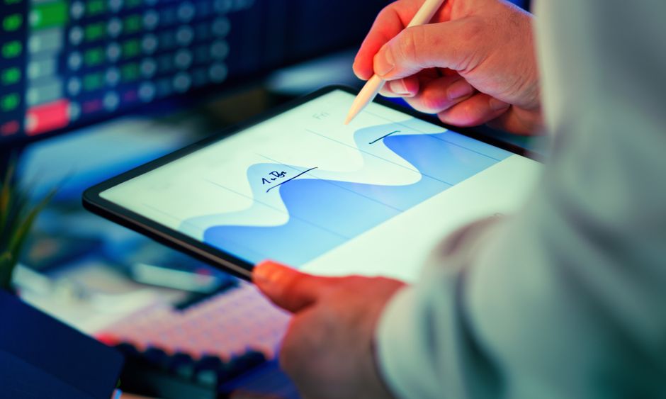 Professionnel analysant un graphique de données sur une tablette numérique avec stylet, écrans de monitoring en arrière-plan