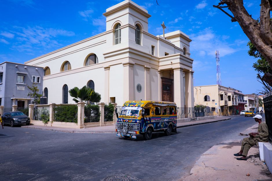 Rue animée de Dakar avec un car rapide coloré devant une église coloniale, symbole de la capitale sénégalaise