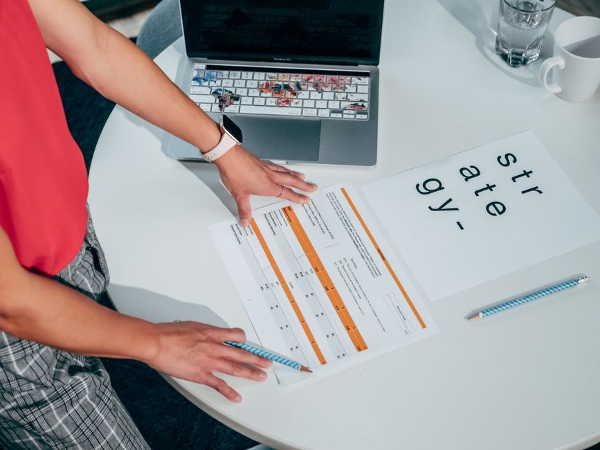Vue aérienne de mains consultant une grille d'évaluation structurée avec colonnes notées sur bureau blanc, laptop en arrière-plan